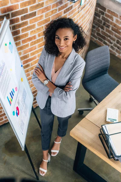 Smiling african american Casual businesswoman with crossed arms near flipchart in loft office — Stock Photo