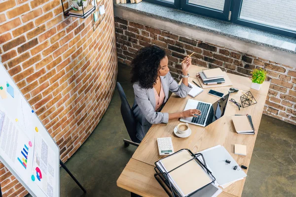 Vista de ángulo alto de la mujer de negocios ocasional afroamericana sentada en el escritorio y usando el ordenador portátil - foto de stock