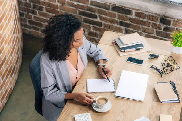 Lässige afrikanisch-amerikanische Geschäftsfrau sitzt am Schreibtisch und schreibt in Notizbuch im Loft-Büro — Stockfoto