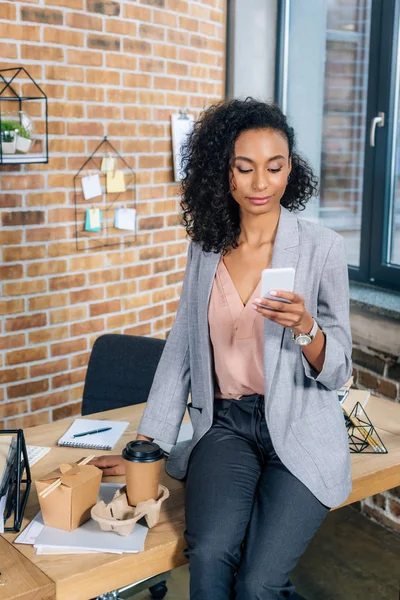 Afrikanisch-amerikanische Gelegenheitsunternehmerin sitzt am Schreibtisch und nutzt Smartphone — Stockfoto