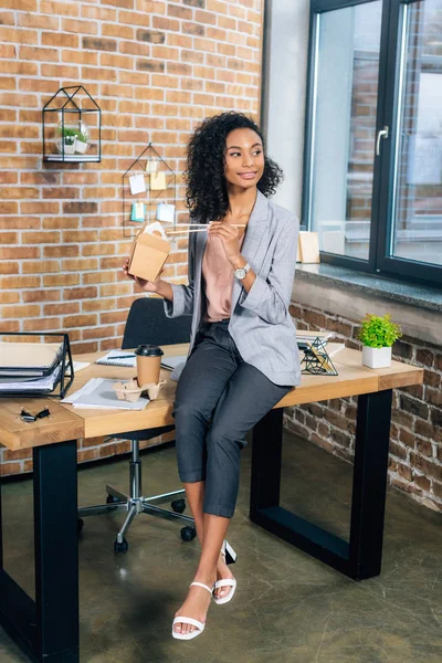 Souriant afro-américain femme d'affaires occasionnelle assis sur le bureau avec du café pour aller et boîte à emporter — Photo de stock