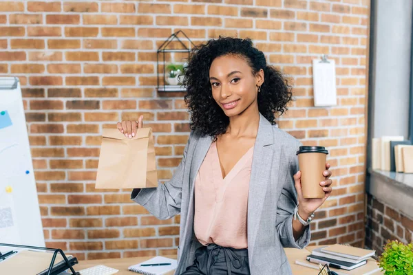 African american Casual businesswoman holding takeaway paper bag and coffee to go in office — Stock Photo