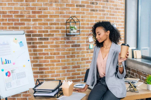 African american Casual businesswoman sitting on desk with coffee to go and takeaway box in office — Stock Photo