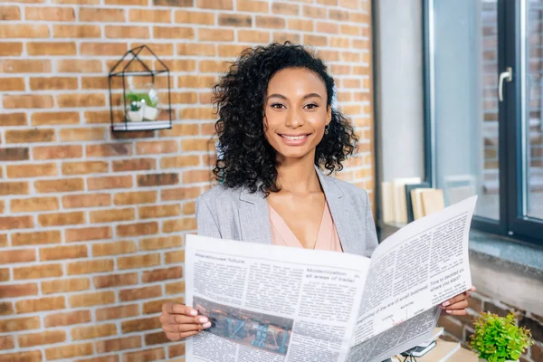 Hermosa sonriente afroamericana ocasional mujer de negocios celebración de periódico de negocios en la oficina loft - foto de stock