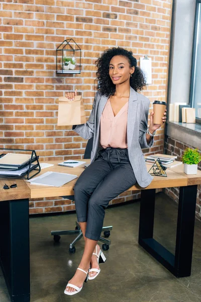 Africano americano casual mujer de negocios holding takeaway papel bolsa y café a ir en oficina - foto de stock