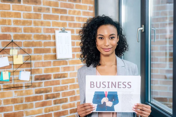 Hermosa afroamericana casual mujer de negocios celebración de periódico de negocios en la oficina loft - foto de stock