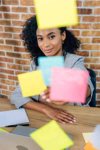 Hermosa mujer de negocios ocasional afroamericana sentada en el escritorio frente a la ventana de vidrio con notas adhesivas - foto de stock