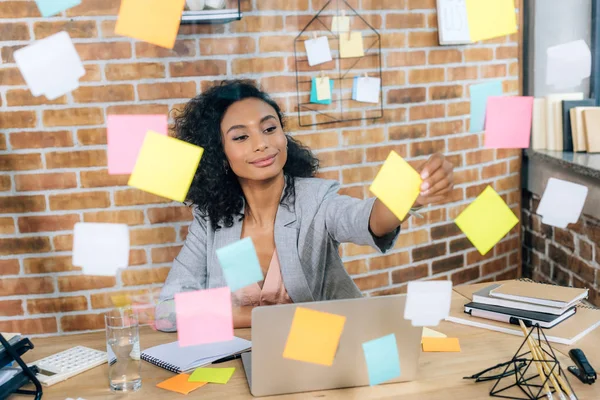 Hermosa afroamericana ocasional mujer de negocios poniendo notas pegajosas en ventana de cristal en la oficina - foto de stock