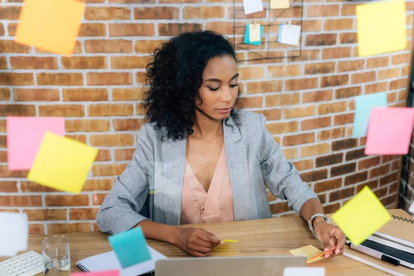 Lässige afrikanisch-amerikanische Geschäftsfrau sitzt am Schreibtisch vor dem Glasfenster mit klebrigen Zetteln — Stockfoto