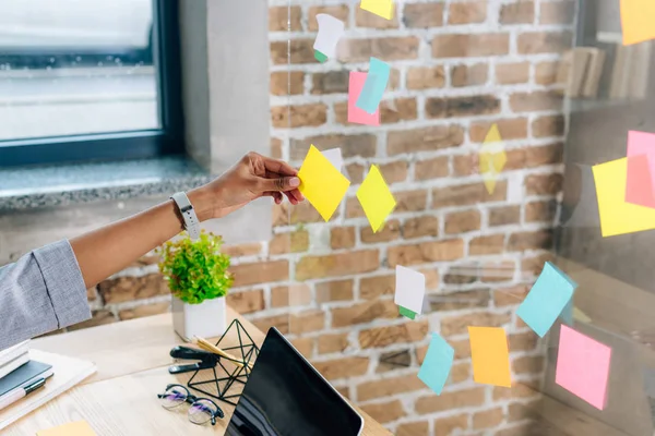 Vue recadrée de femme afro-américaine mettant Sticky Notes sur fenêtre en verre dans le bureau loft — Photo de stock