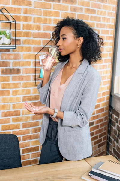 African american Casual businesswoman with pills drinking water in office — Stock Photo