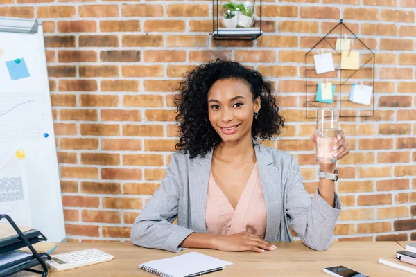 Smiling african american Casual businesswoman holding glass of water at office desk — Stock Photo