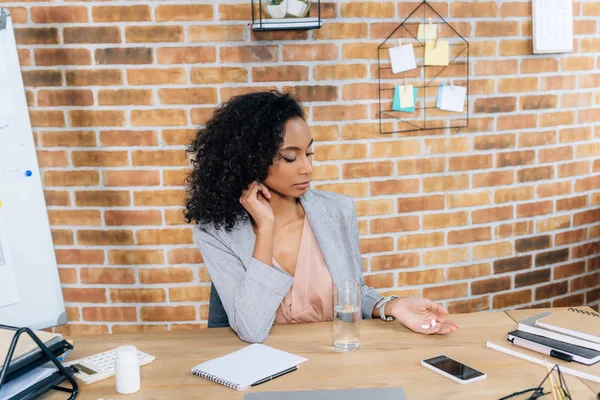 African american Casual businesswoman with pills and glass of water at office desk — Stock Photo