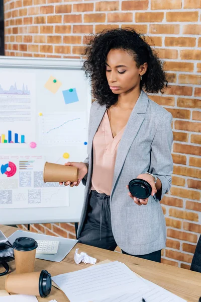 Africano americano casual mujer de negocios celebración de café para ir cerca de flipchart en loft oficina - foto de stock