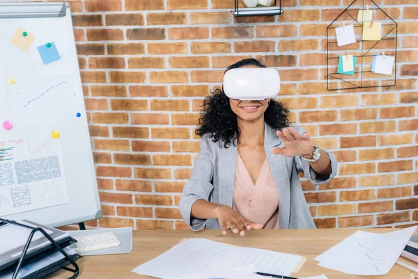 African american Casual businesswoman in virtual reality headset gesturing and sitting at office desk — Stock Photo