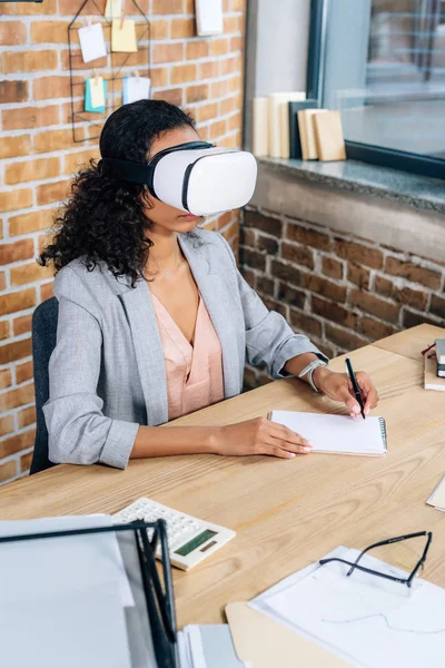 African american Casual businesswoman in virtual reality headset writing in notebook at office desk — Stock Photo