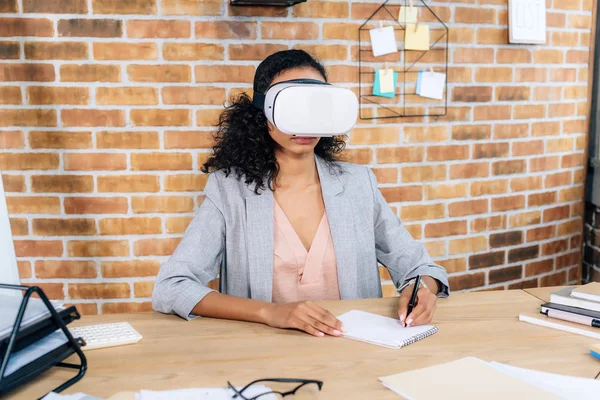 African american Casual businesswoman in virtual reality headset at office desk writing in notebook — Stock Photo