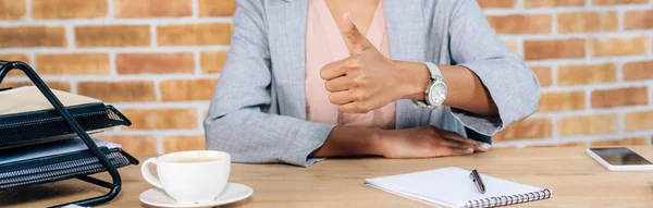 Panoramic shot of Casual african american businesswoman at desk with coffee and notebook showing thumb up — Stock Photo