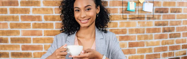 Tiro panorâmico de sorridente afro-americano Casual empresária com xícara de café no escritório — Fotografia de Stock