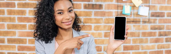 Panoramaaufnahme einer afrikanisch-amerikanischen Geschäftsfrau, die mit dem Finger auf ein Smartphone mit leerem Bildschirm zeigt — Stockfoto