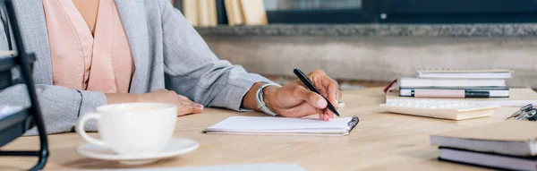 Plano panorámico de la mujer de negocios ocasional afroamericana escribiendo en un cuaderno en el escritorio - foto de stock