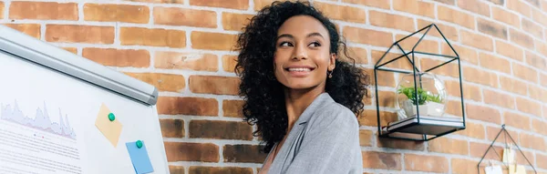 Plan panoramique de femme d'affaires afro-américaine occasionnelle près de tableau à feuilles mobiles dans le bureau loft — Photo de stock