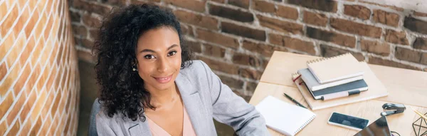 Panoramaaufnahme einer afrikanisch-amerikanischen Geschäftsfrau, die am Schreibtisch im Loft-Büro in die Kamera blickt — Stockfoto