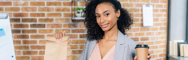 Plano panorámico de la mujer de negocios ocasional afroamericana sosteniendo la bolsa de papel para llevar y café para ir en la oficina - foto de stock