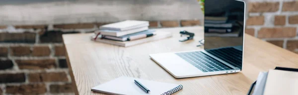 Panoramic shot of wooden desk with laptop, notebook and pen in loft office — Stock Photo