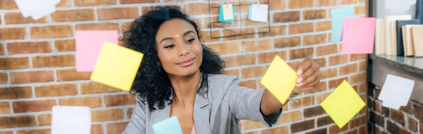 Plan panoramique de femme d'affaires afro-américaine occasionnelle mettant Sticky Notes sur fenêtre en verre dans le bureau — Photo de stock