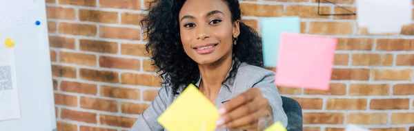 Panoramaaufnahme einer afrikanisch-amerikanischen Geschäftsfrau, die im Büro klebrige Zettel an Glasfenster klebt — Stockfoto