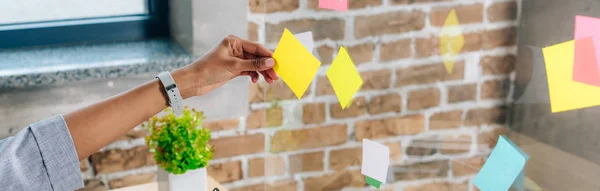 Panoramic shot of african american woman putting Sticky Notes on glass window in loft office — Stock Photo