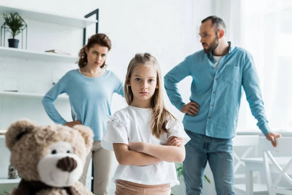 Enfoque selectivo de niño lindo de pie con brazos cruzados cerca de oso de peluche y los padres en casa - foto de stock