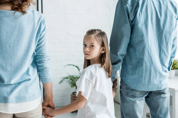 Грустный ребенок, стоящий с родителями и держащийся за руки дома — стоковое фото