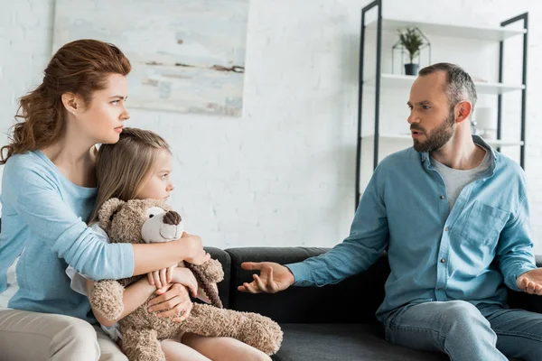 Mother hugging daughter with teddy bear and looking at upset husband at home — Stock Photo