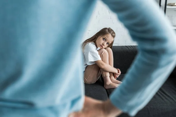 Selective focus of scared child sitting on sofa at looking at mother standing with hand on hip — Stock Photo