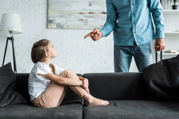 Cropped view of man pointing with finger at upset kid sitting on sofa at home — Stock Photo