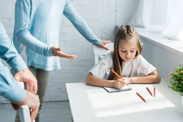 Cropped view of parents standing near upset daughter holding pencil at home — Stock Photo