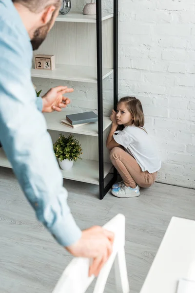 Selektiver Fokus des verängstigten Kindes, das den Vater ansieht, während es in der Nähe des Regals zu Hause sitzt — Stockfoto