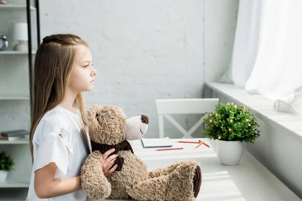 Niedliches Kind hält Teddybär, während es am Tisch zu Hause steht — Stockfoto