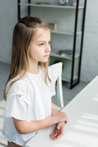 Adorable niño en camiseta blanca de pie cerca de la mesa en casa - foto de stock