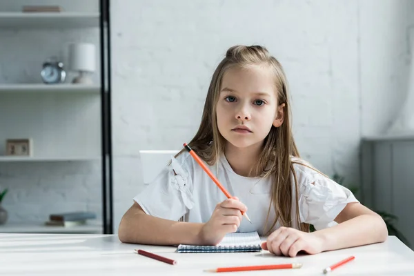 Cute kid holding pencil near notebook while looking at camera at home — Stock Photo