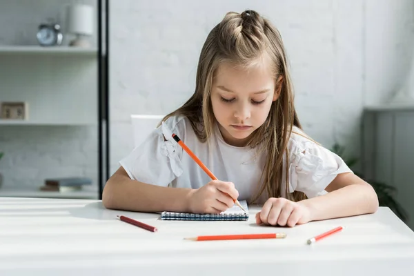 Niedliches Kind hält Bleistift beim Schreiben in Notizbuch zu Hause — Stockfoto