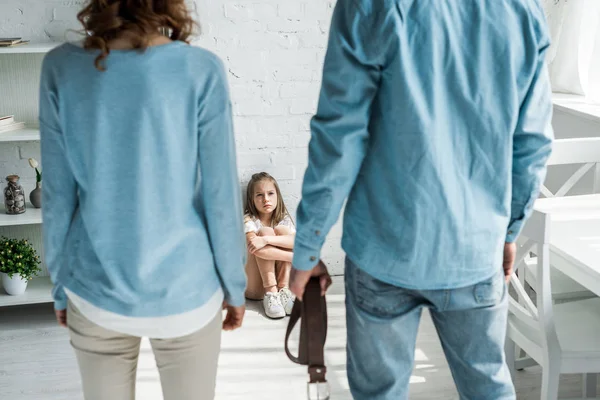 Избирательный фокус испуганного ребенка, сидящего на полу и смотрящего на отца с ремнем, стоящего рядом с матерью — стоковое фото