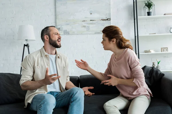 Upset woman gesturing and looking at bearded man talking while sitting on sofa — Stock Photo