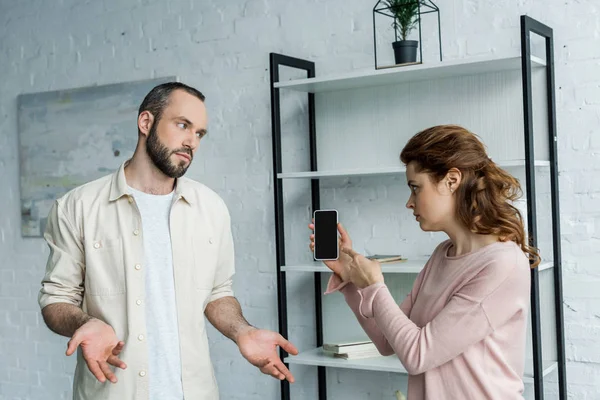 Mujer ofendida señalando con el dedo en el teléfono inteligente con la pantalla en blanco cerca del hombre en casa - foto de stock