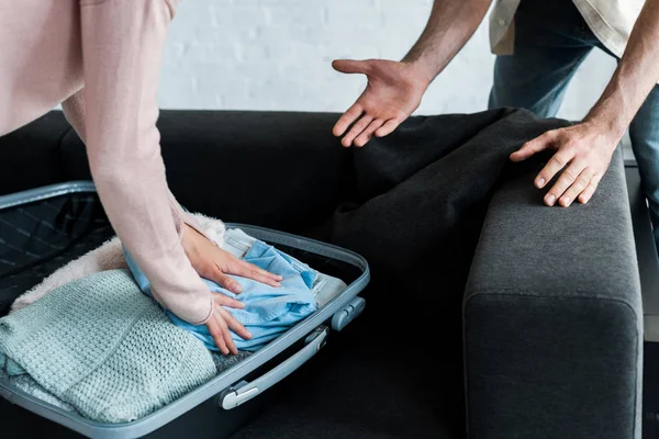 Cropped view of man gesturing near woman standing and packing clothes in suitcase — Stock Photo