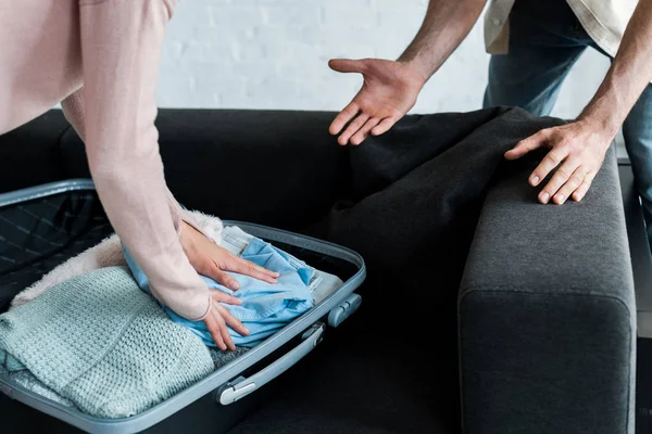 Recortado vista de hombre gesto cerca de mujer de pie y embalaje de ropa en maleta - foto de stock