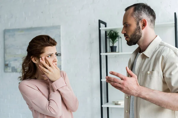 Verängstigte Frau berührt Gesicht und sieht wütenden Mann zu Hause an — Stockfoto