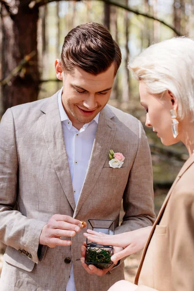 Vista lateral del hombre sonriente poniéndose el anillo de bodas en el dedo de la mujer en el bosque - foto de stock